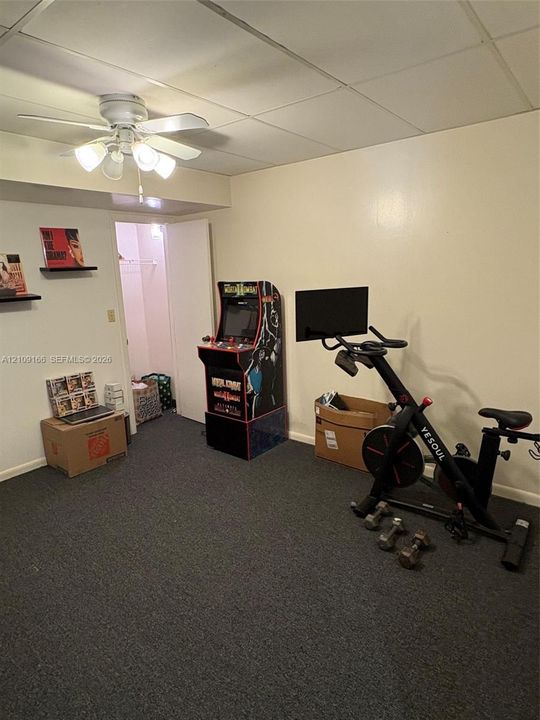 Neutral-toned interior space featuring dark wall-to-wall carpeting, a drop ceiling, a ceiling-mounted light fixture with four glass shades, and an enclosed storage closet
