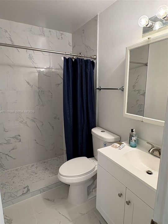 Bathroom featuring shower walls and flooring finished in white stone-pattern tile, white vanity cabinet with integrated basin, polished metal faucet, and wall-mounted medicine cabinet with integrated lighting