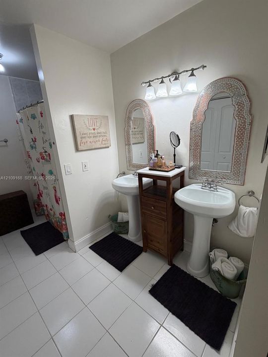 Dual white pedestal sinks with polished chrome fixtures, complemented by wall-mounted mirrors and four-light vanity lighting over ceramic tile flooring