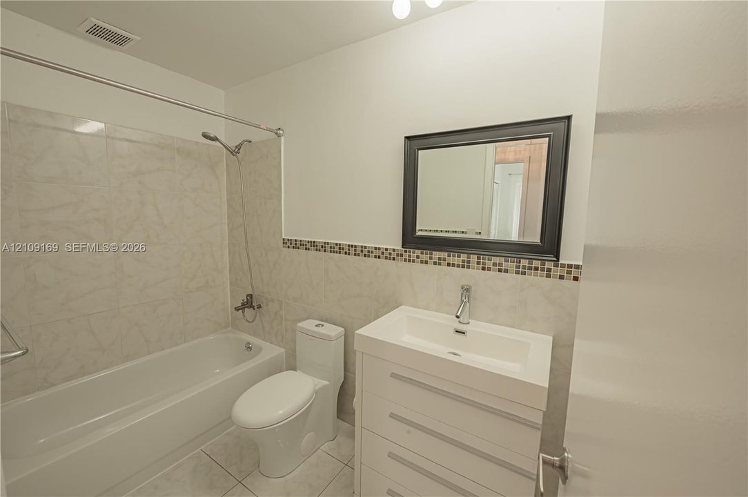Bathroom featuring a white vanity cabinet with a rectangular integrated sink, neutral stone-finish tile wainscoting with a glass mosaic accent border, and a shower-tub combination with matching wall tiling