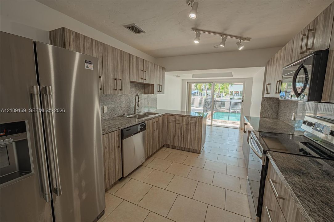 Kitchen featuring wood-finish cabinetry, stone-surface countertops, stainless steel appliances, ceramic tile flooring, and adjustable track lighting