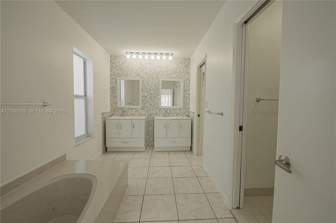 Dual white vanities, multi-colored stone-tiled accent wall, built-in soaking tub, large-format tile flooring, and multi-bulb integrated vanity lighting