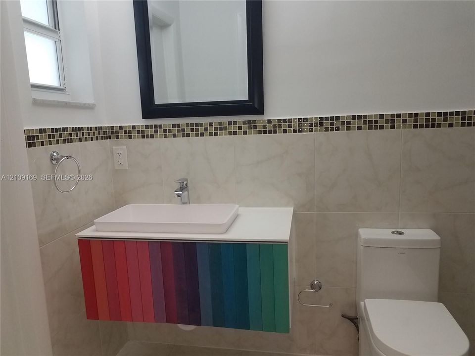 Wall-mounted floating vanity with a rectangular vessel sink and multi-colored paneling, complemented by a mosaic tile backsplash, framed wall mirror, and dual-flush toilet