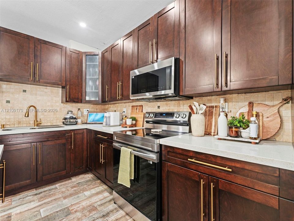 Kitchen featuring dark wood cabinetry, light-toned stone-style countertops, mosaic tile backsplash, stainless steel appliances, and a brass-tone gooseneck faucet