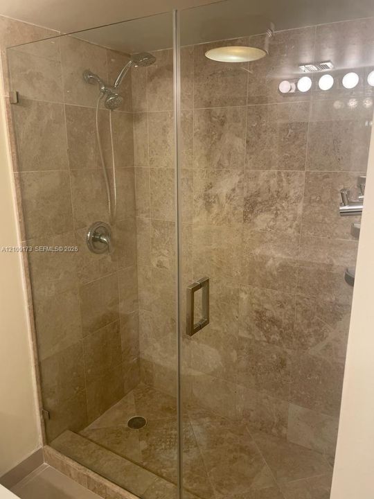 Walk-in shower featuring floor-to-ceiling stone tile, a frameless glass enclosure, a polished metal handheld shower head, and a recessed ceiling light