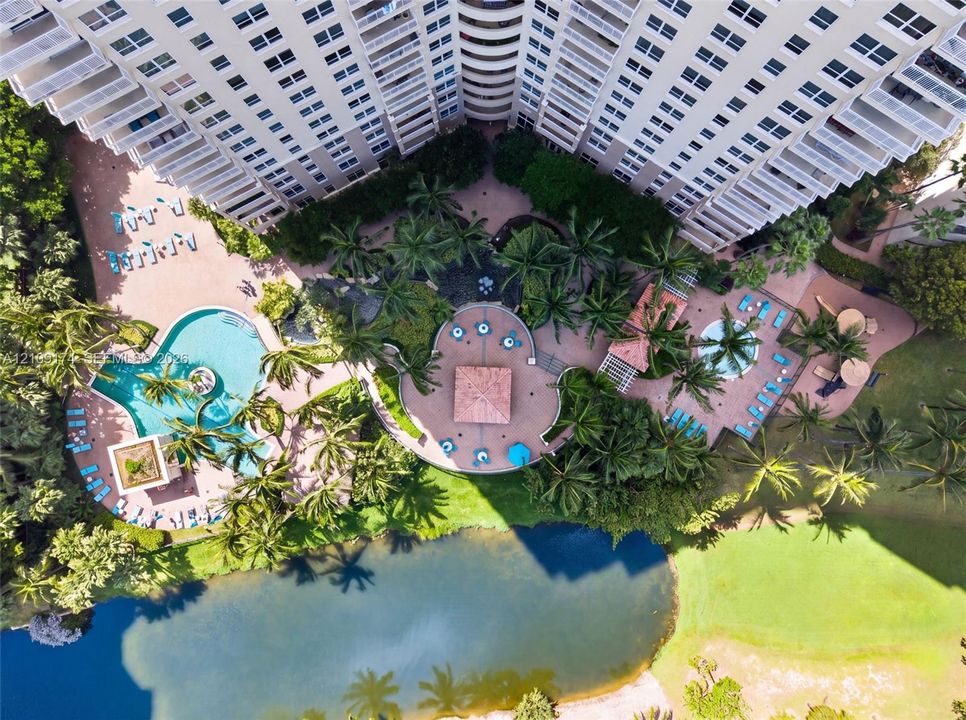 Aerial perspective featuring a multi-story high-rise building with wraparound balconies, an expansive swimming pool, a separate circular spa, a paved central plaza with a tile-roofed gazebo, and manicured landscaping bordering a water feature