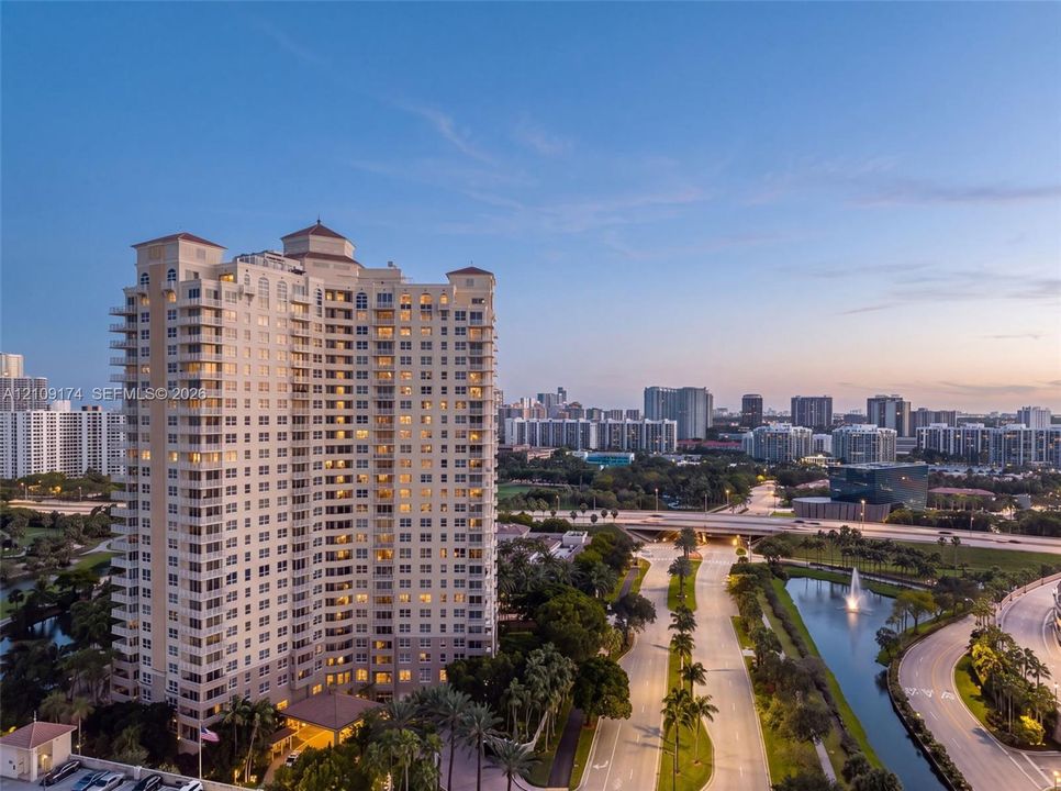 High-rise residential tower featuring a tiered architectural design, private balconies, a covered porte-cochère entry, mature palm tree landscaping, and a surrounding water feature