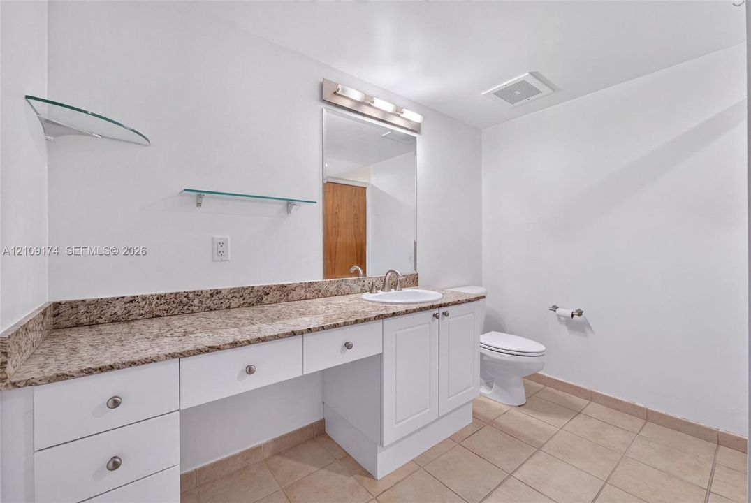 White cabinetry with stone-finish countertop, undermount sink, integrated vanity lighting, glass shelving, and ceramic tile flooring