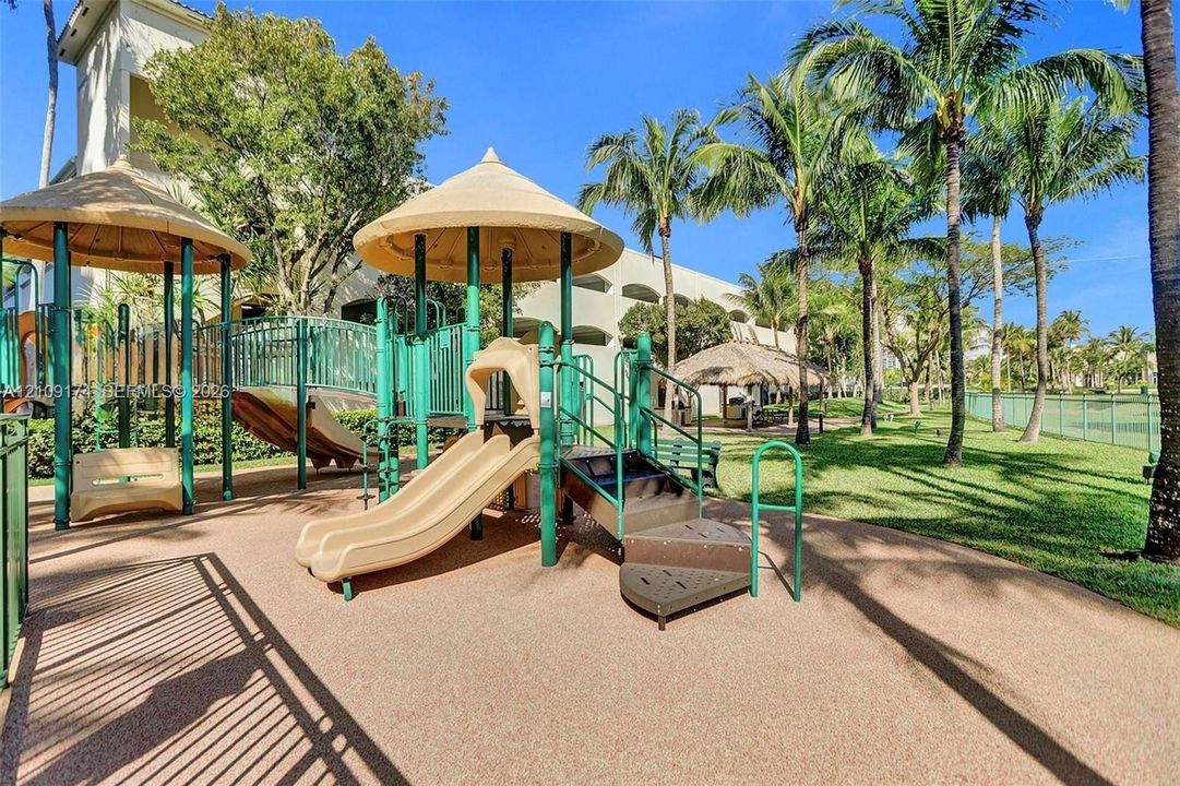Recreational play area featuring a multi-level structure with slides, shaded canopies, and a paved rubberized surface, set within a landscaped perimeter with mature palm trees
