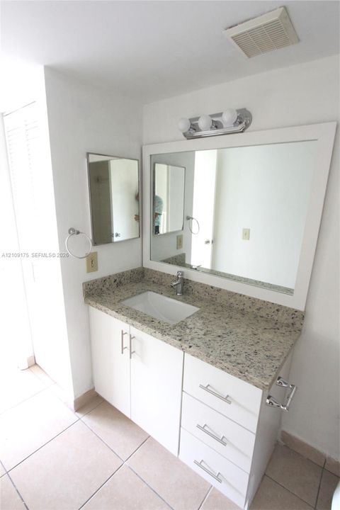Bathroom vanity featuring stone-finish countertop, undermount sink, white cabinetry with silver-tone hardware, and a framed mirror with a three-bulb light fixture