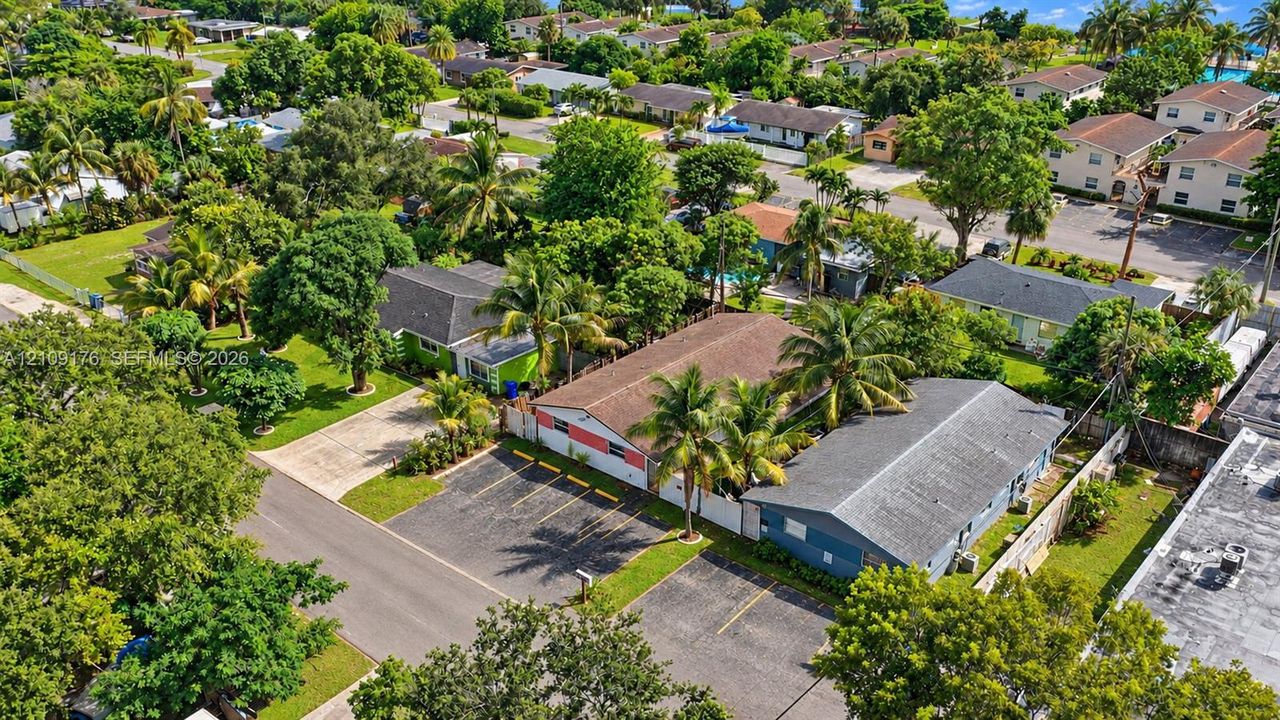 Aerial perspective of the property featuring a paved off-street parking area, multi-tone exterior siding, and a shingled roof, surrounded by mature palm trees and established landscaping
