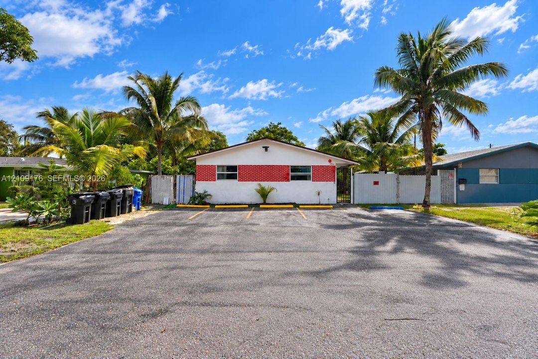 Exterior featuring a white facade with red masonry accents, a paved parking area with designated spaces, and mature palm trees