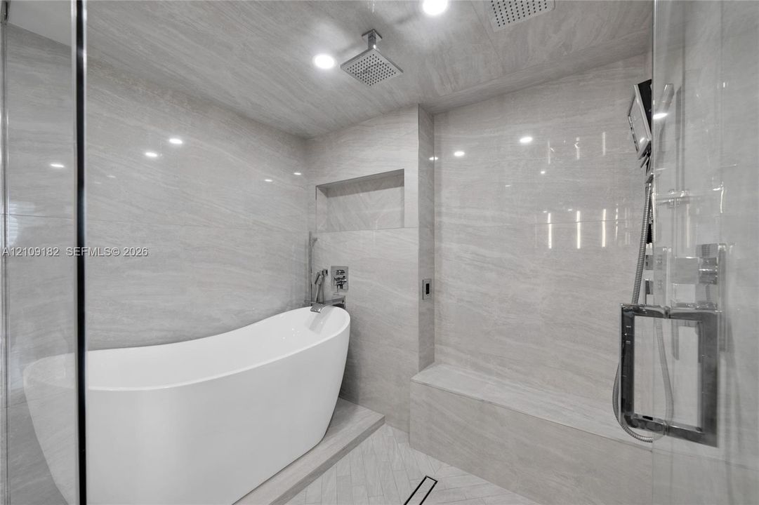 Freestanding soaking tub, floor-to-ceiling stone-finish tile surround, built-in recessed shower niche, integrated stone bench, and glass-enclosed shower featuring dual showerheads