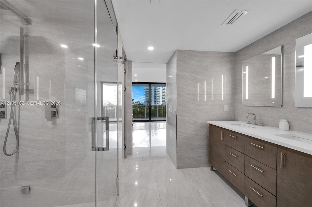 Double vanity with wood-finish cabinetry, white countertop, integrated dual sinks, and frameless glass-enclosed shower with tiled wall surfaces