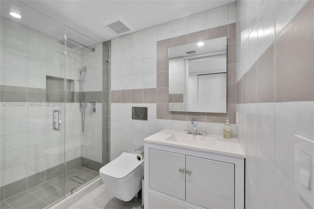Bathroom featuring a walk-in glass-enclosed shower, wall-mounted dual-flush toilet, vanity with integrated basin, polished chrome fixtures, and neutral-toned wall tile
