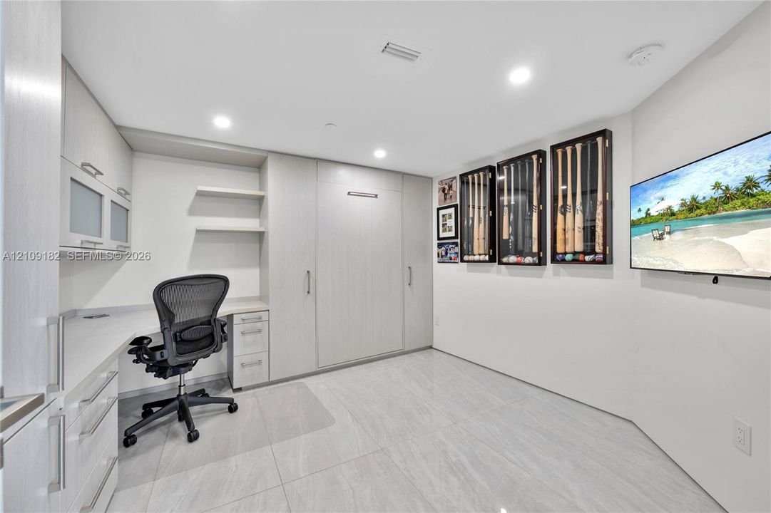 Custom built-in cabinetry featuring a desk station, integrated shelving, and a wall-mounted murphy bed unit, all finished in a light wood-grain veneer, complemented by large-format porcelain tile flooring and recessed LED lighting