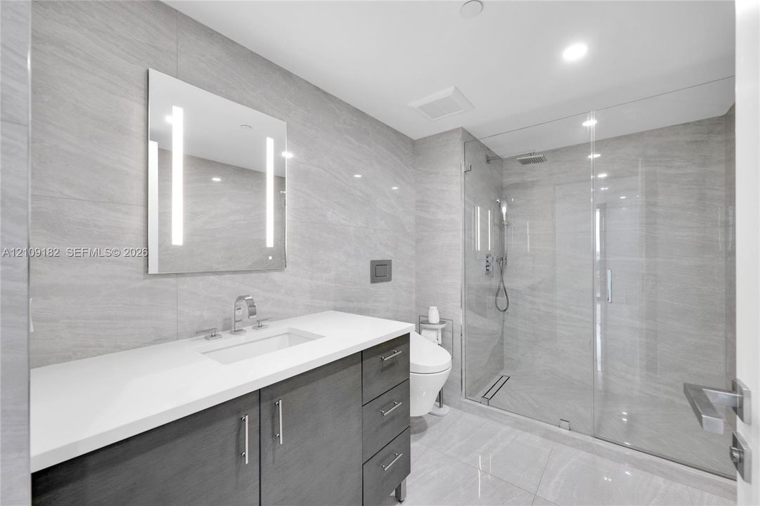 Modern vanity with stone countertop and integrated sink, full-height stone wall tiling, LED-backlit mirror, glass-enclosed shower with rainfall showerhead, and wall-mounted toilet