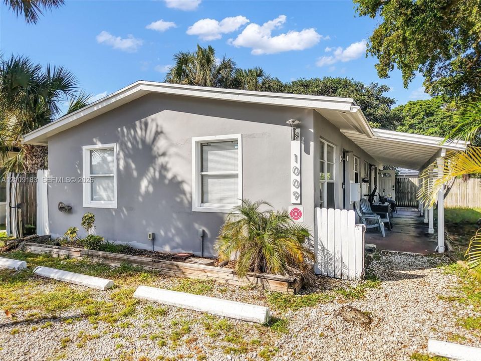 Exterior featuring a light grey stucco finish, a covered concrete patio, white picket fencing, and a gravel parking area with concrete wheel stops