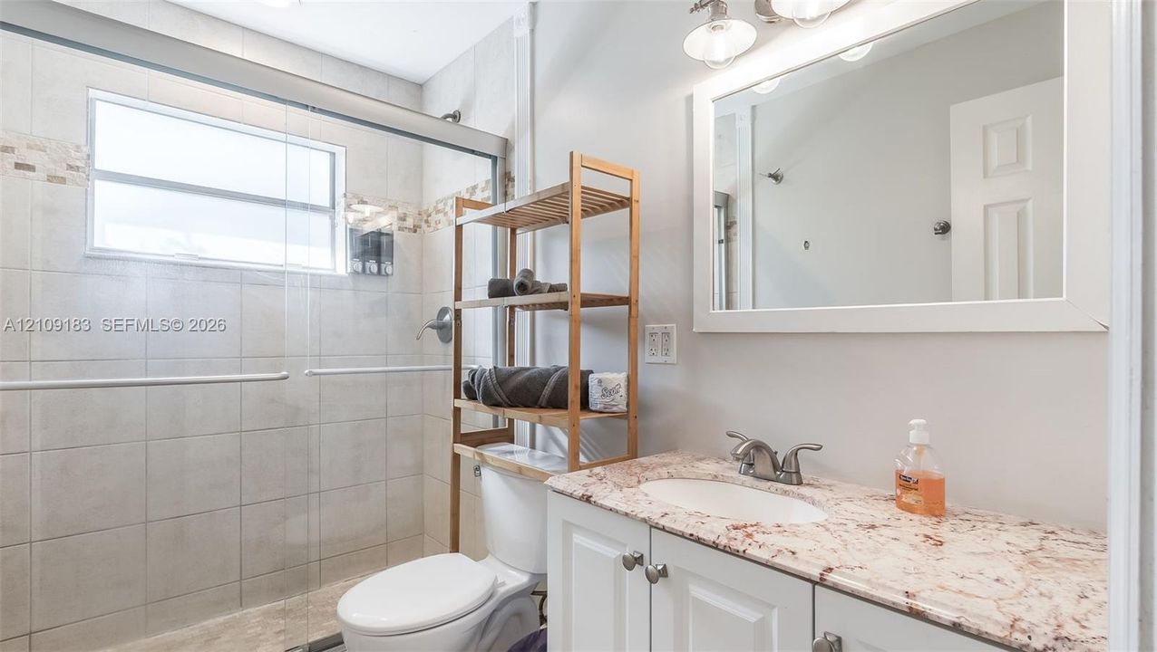 Bathroom featuring a stone-surface vanity with an integrated sink, white cabinetry, a glass-enclosed shower with tiled walls, and a wall-mounted vanity mirror with integrated lighting