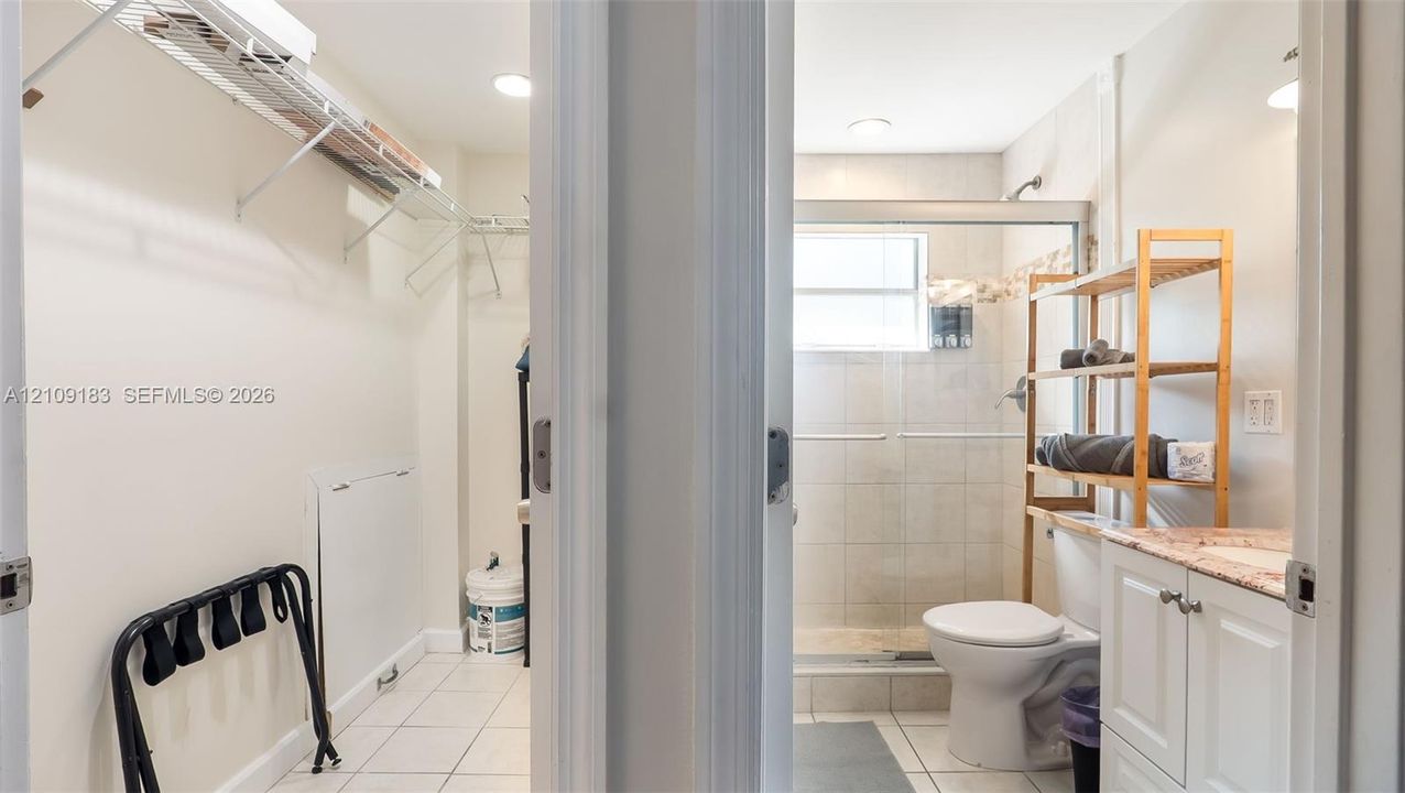 Storage area featuring wire shelving and white tile flooring; bathroom including a vanity with a light-toned stone countertop, a walk-in shower with tiled surround, and recessed lighting
