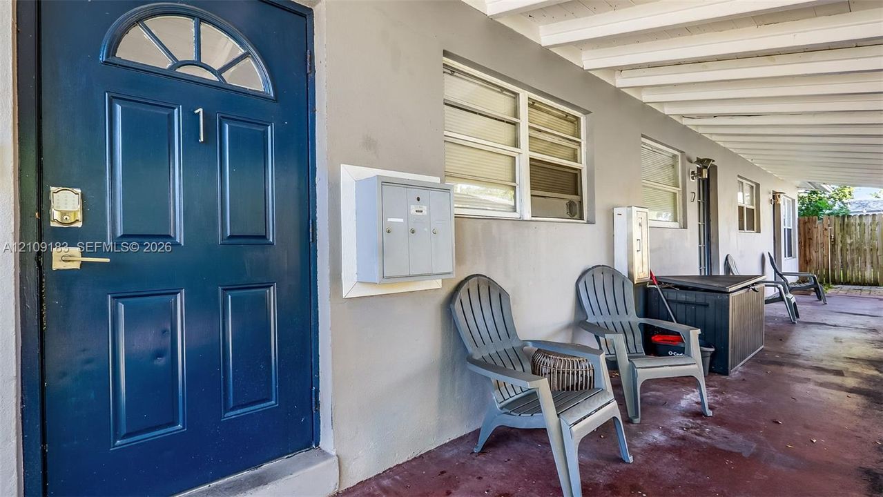 Covered exterior walkway featuring a solid-panel door with an arched window inlay, horizontal sliding windows, and a wall-mounted metal mailbox