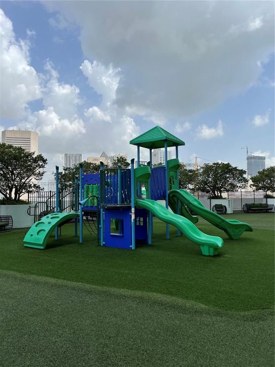 Recreational area featuring a multi-component play structure with dual slides and climbing elements, set upon a synthetic grass surface and enclosed by perimeter fencing