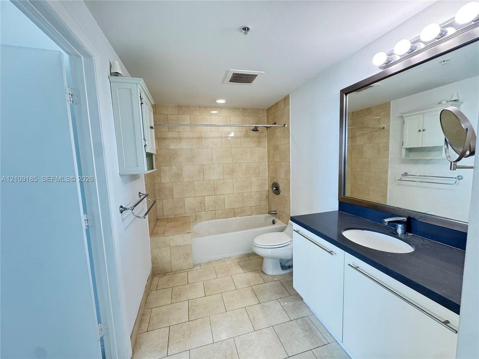 Bathroom featuring white cabinetry, dark countertop with an integrated oval sink, full-width mirror with integrated lighting, and a tile-surround shower and tub combination