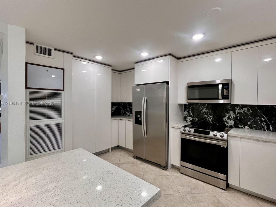 Kitchen featuring sleek white flat-panel cabinetry, light-toned stone-finish countertops, stainless steel appliances, and a high-contrast dark stone backsplash