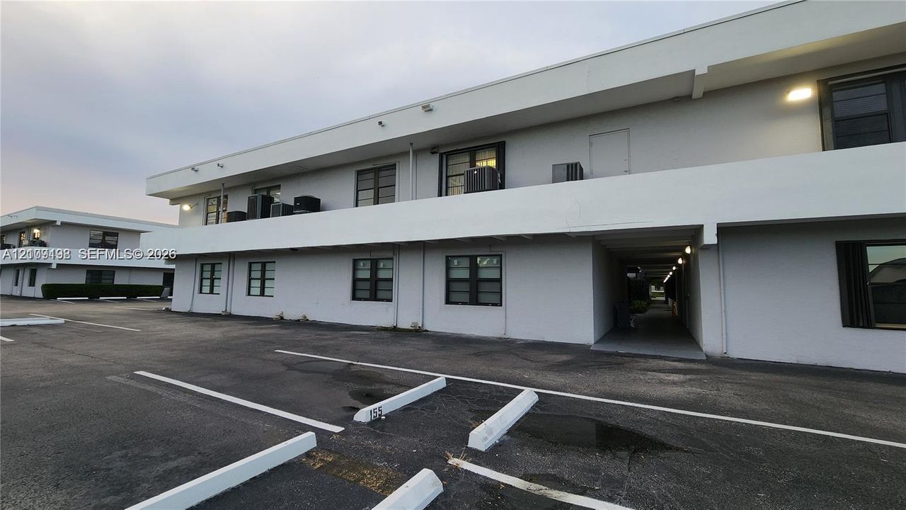 Low-rise concrete exterior featuring multi-pane windows, covered walkways, and asphalt surface parking with designated wheel stops