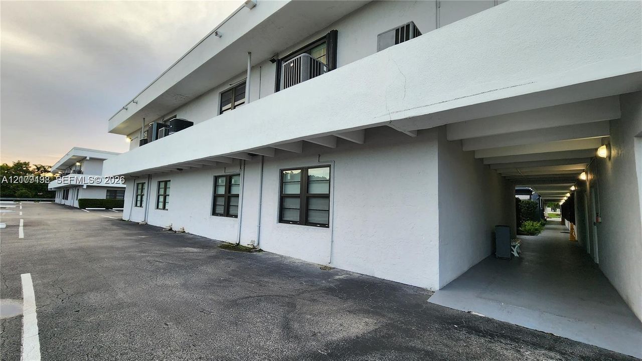 Multi-story exterior featuring white stucco siding, dark-framed casement windows, a covered ground-level walkway, and an expansive paved parking area