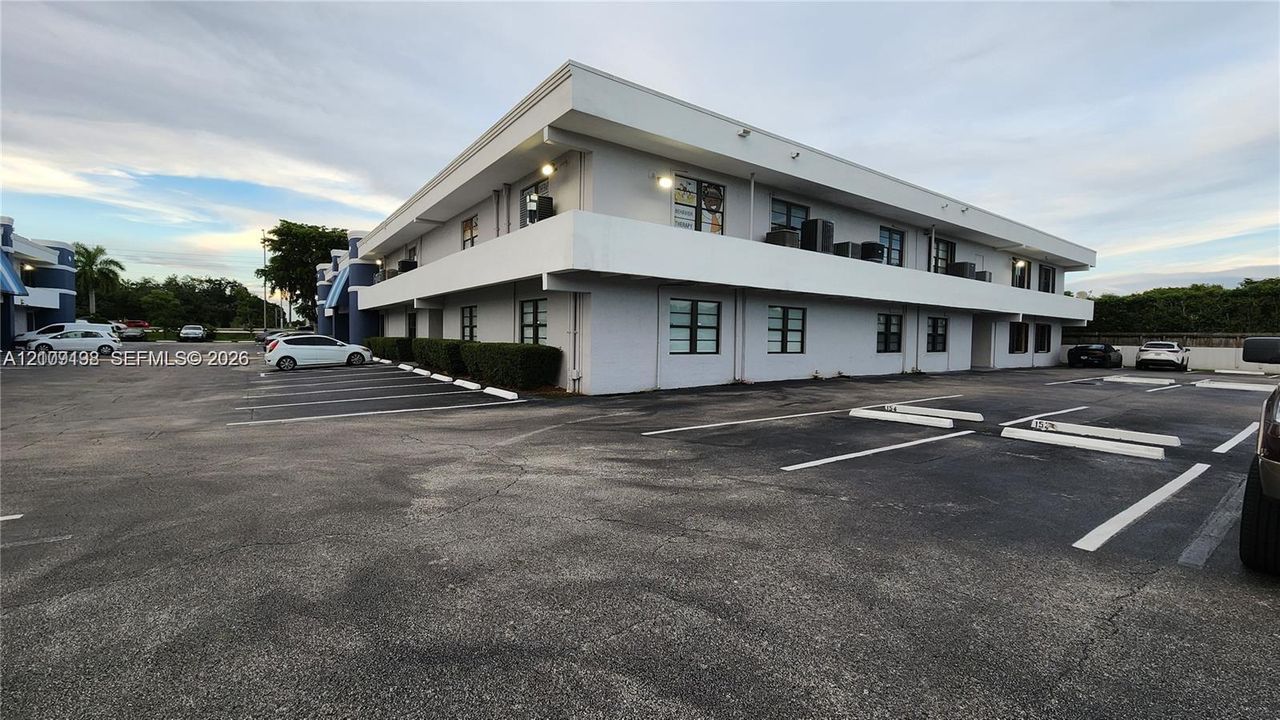 Two-story commercial exterior featuring a flat roofline, second-level balcony, painted masonry facade, and an asphalt parking lot with designated spaces