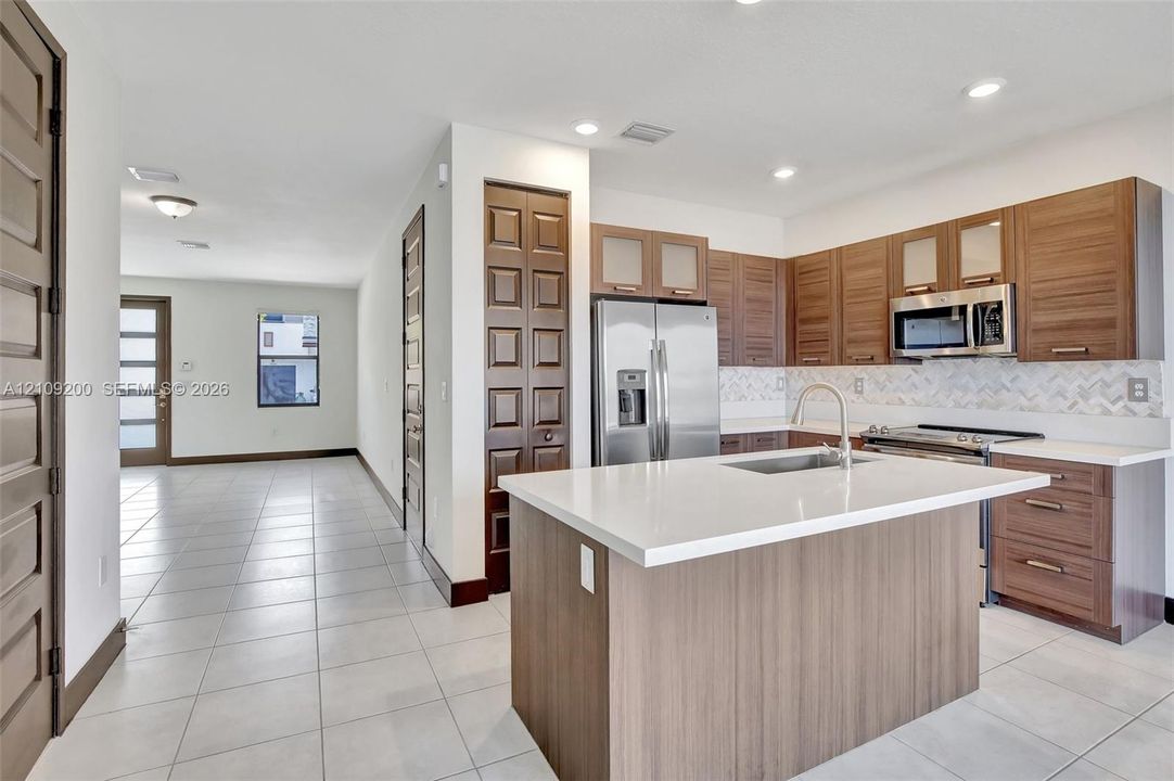 Kitchen featuring wood-finish cabinetry, a central island with a solid-surface countertop, stainless steel appliances, a chevron-pattern backsplash, and tile flooring