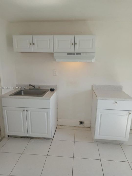 Kitchen area featuring white cabinetry, light-toned composite countertops, a stainless steel sink with a dual-handle faucet, a mounted range hood, and white tile flooring