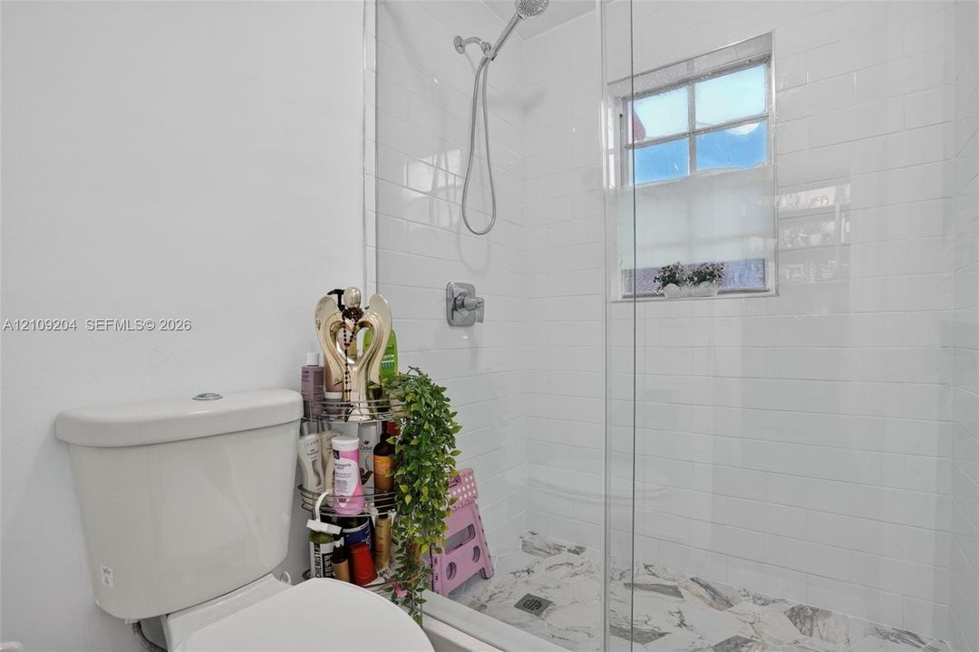 Walk-in shower featuring white subway tile surround, frameless glass door, handheld shower fixture, and patterned stone-finish flooring with a multi-pane window