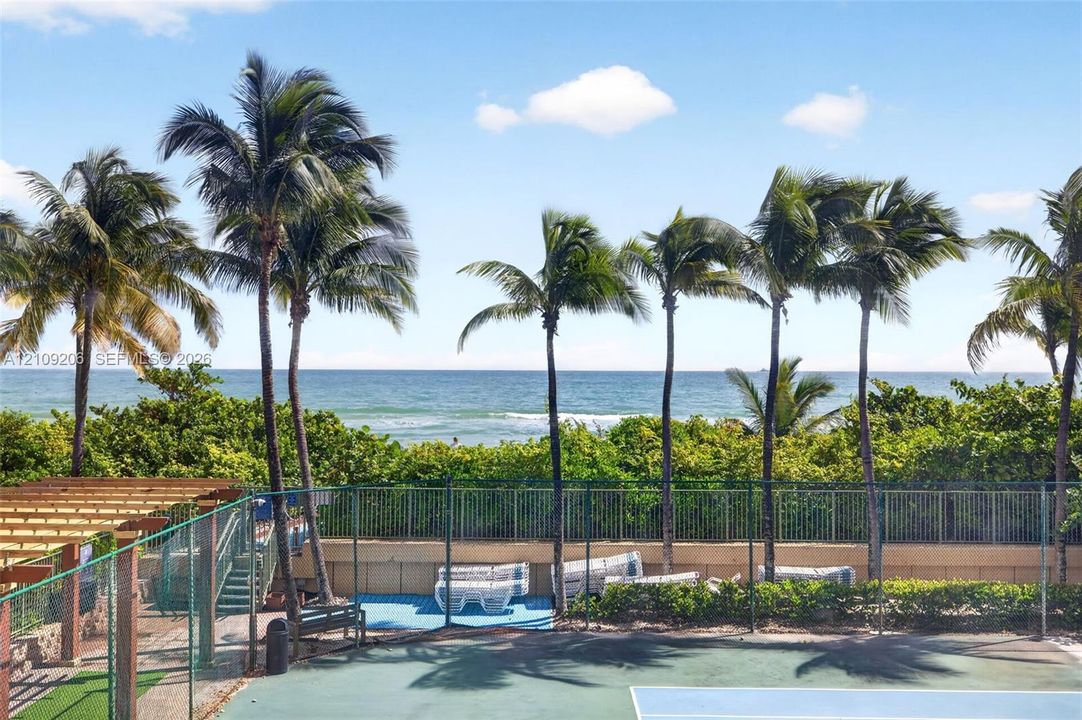 Tennis court enclosed by perimeter fencing, featuring a wooden pergola, mature palm trees, and ocean frontage
