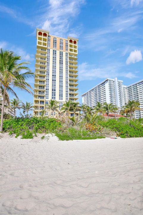 High-rise structure featuring stacked balconies, exterior arch details, sand-covered beach, and lush palm-lined vegetation