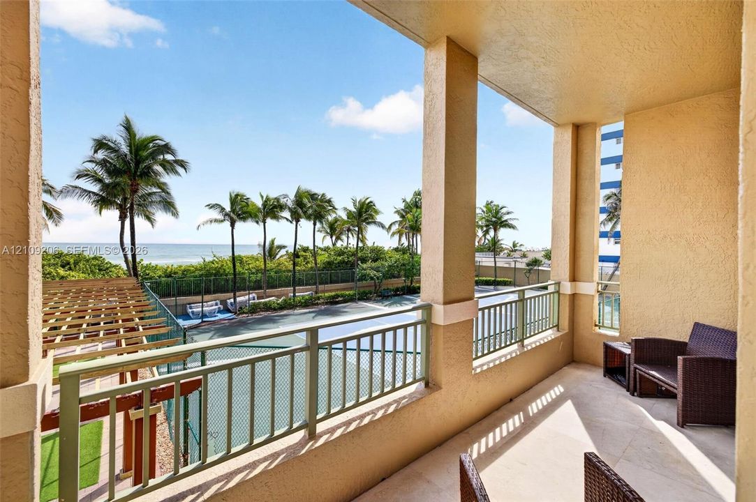 Covered balcony featuring stone-finish flooring, metal railing, and structural pillars, overlooking a hard-surface court and tropical landscaping