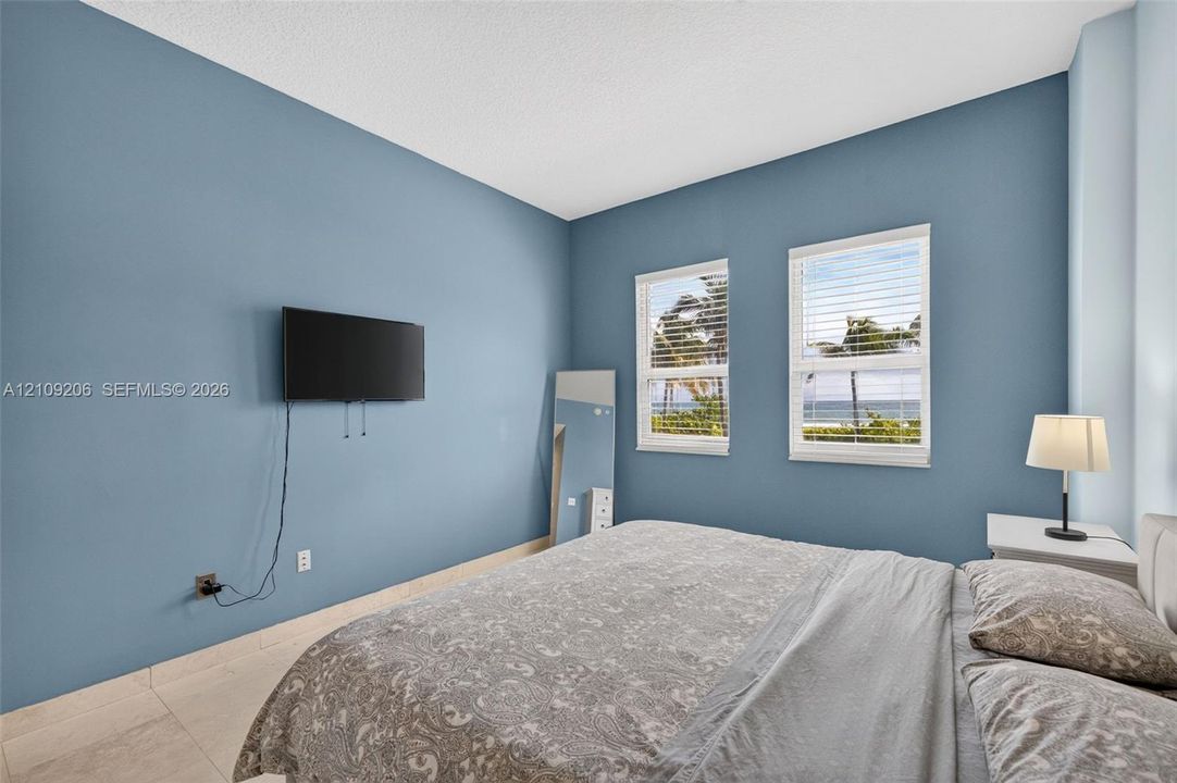 Spacious bedroom featuring light-toned tile flooring, two windows with horizontal blinds, and a coastal landscape visible through the glass