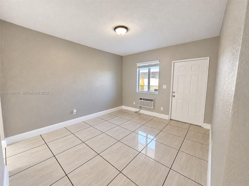 Neutral-toned room featuring large-format rectangular floor tiling, a single-window wall, a wall-mounted air conditioning unit, and a six-panel exterior door