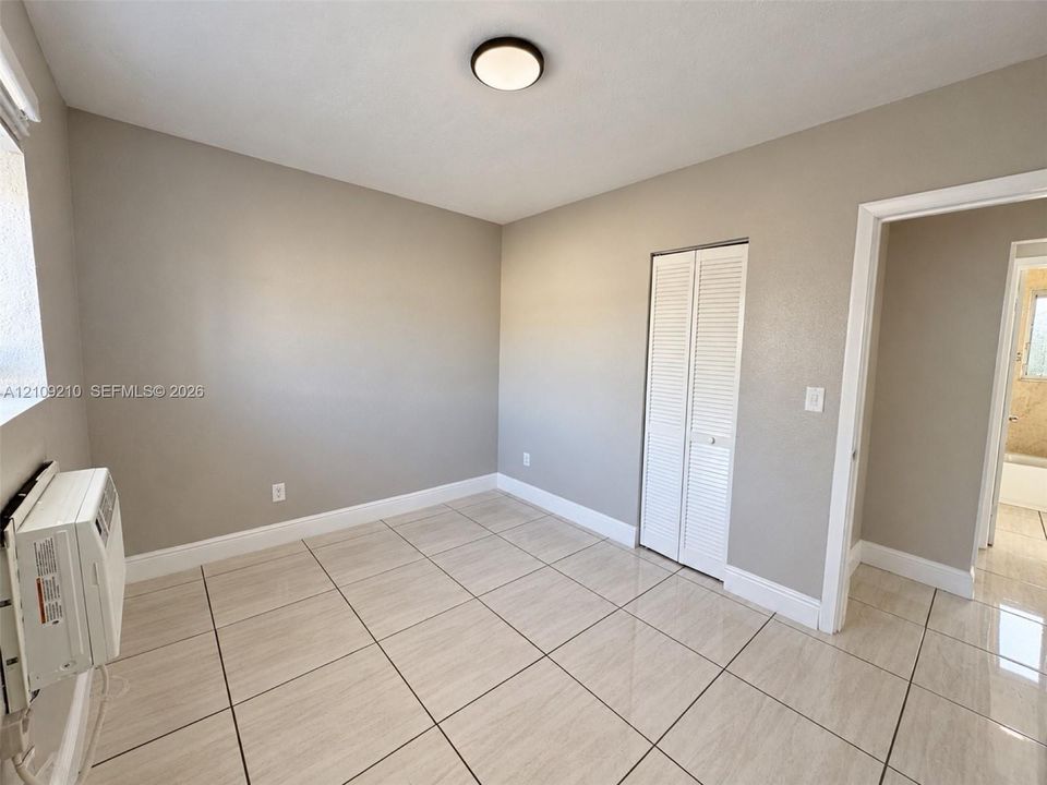 Room featuring neutral wall paint, large-format tile flooring, a dual-panel closet with louvered doors, and a wall-mounted climate control unit