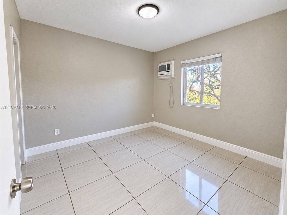 Room featuring large-format neutral tile flooring, smooth wall finishes, a window with horizontal blinds, and a built-in wall-mounted air conditioning unit