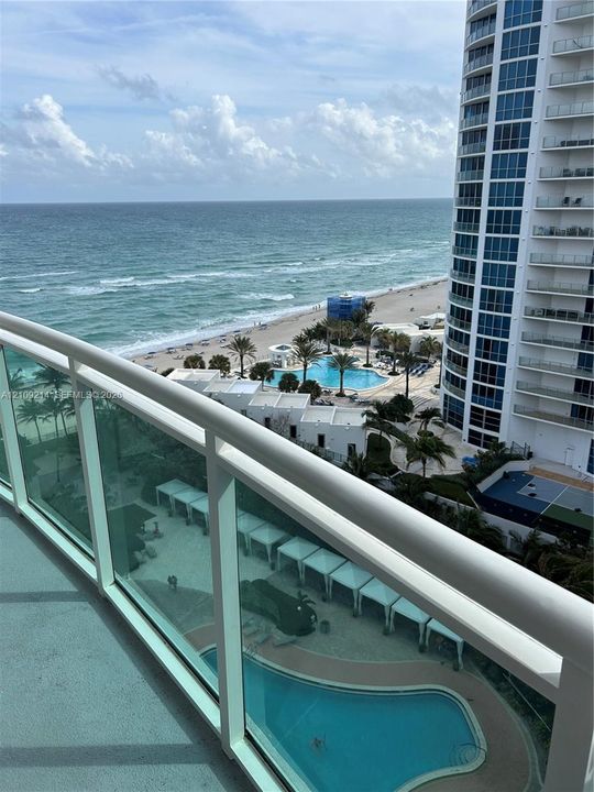 Private balcony featuring glass railings, overlooking a swimming pool, cabanas, and oceanfront shoreline