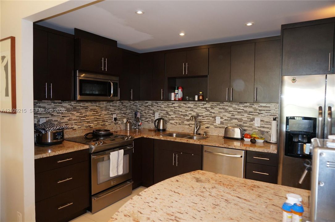Kitchen featuring dark-toned cabinetry, light-toned stone countertops, multi-tonal mosaic tile backsplash, recessed lighting, and stainless steel appliances