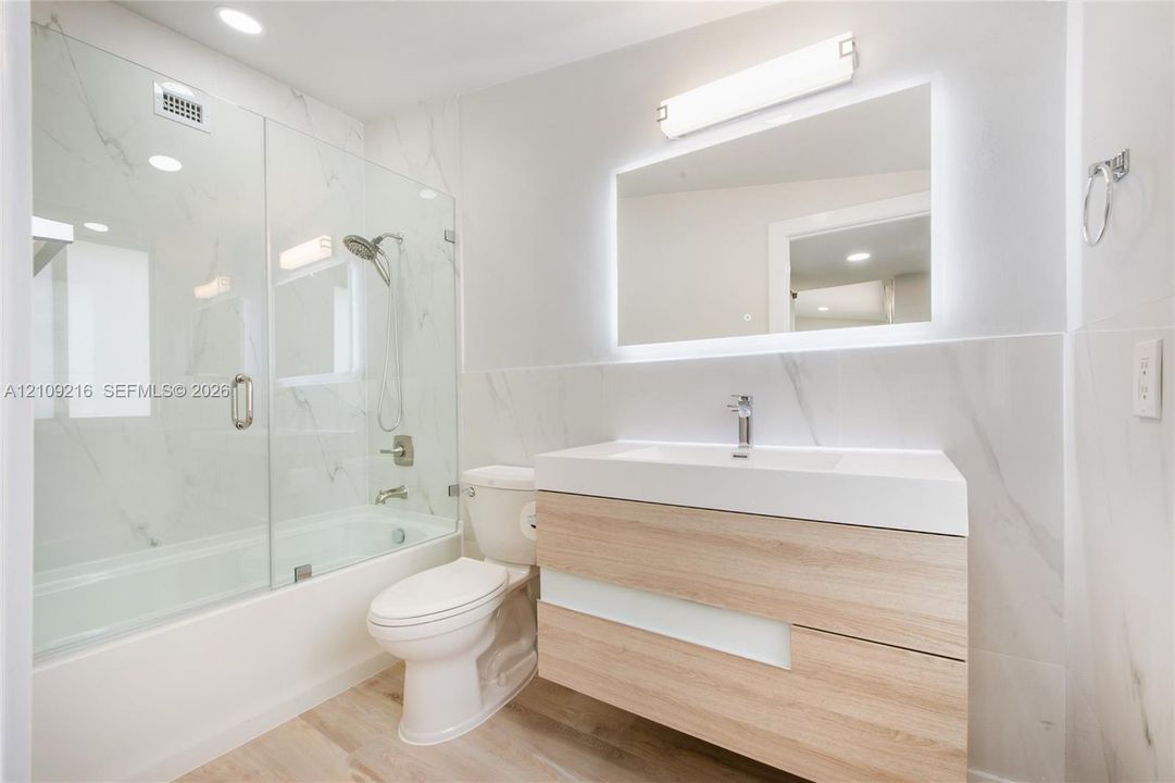 Modern floating vanity with a white integrated sink, wood-finish cabinetry, illuminated wall mirror, glass-enclosed shower and tub combination, and large-format tile surround