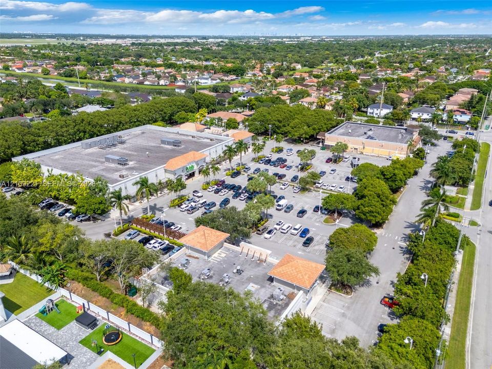 Aerial view of a commercial plaza featuring a flat-roofed building, expansive asphalt parking area, palm trees, and terracotta-style tile roof accents