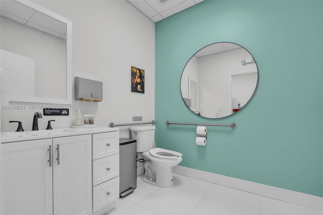 Restroom featuring white cabinetry with brushed metal hardware, a white vanity top with a dark-finish faucet, a large circular wall mirror, and metal wall-mounted grab bars