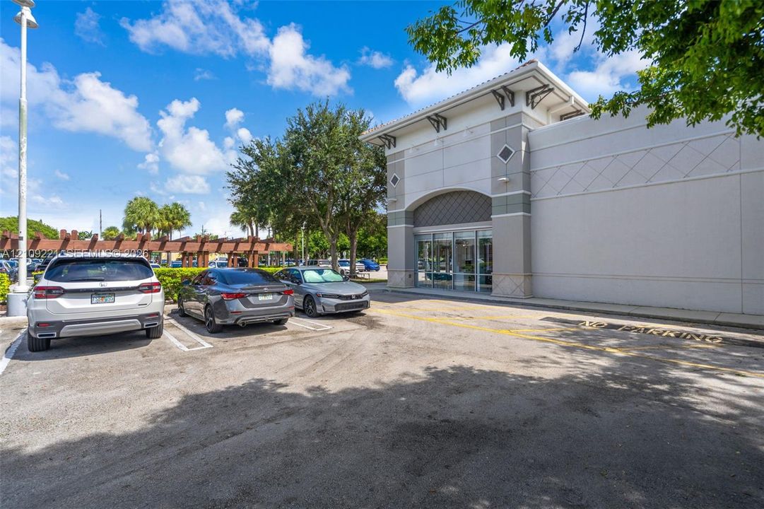 Commercial exterior featuring a stucco facade, decorative corbel accents, a glass storefront entry, and an attached pergola structure over a paved parking area