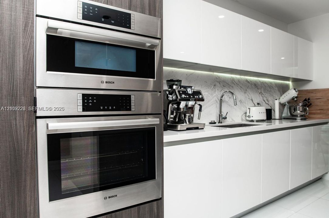 Modern kitchen featuring a stacked stainless steel Bosch wall oven and microwave, high-gloss white cabinetry, stone-finish backsplash, and an undermount sink with a gooseneck faucet