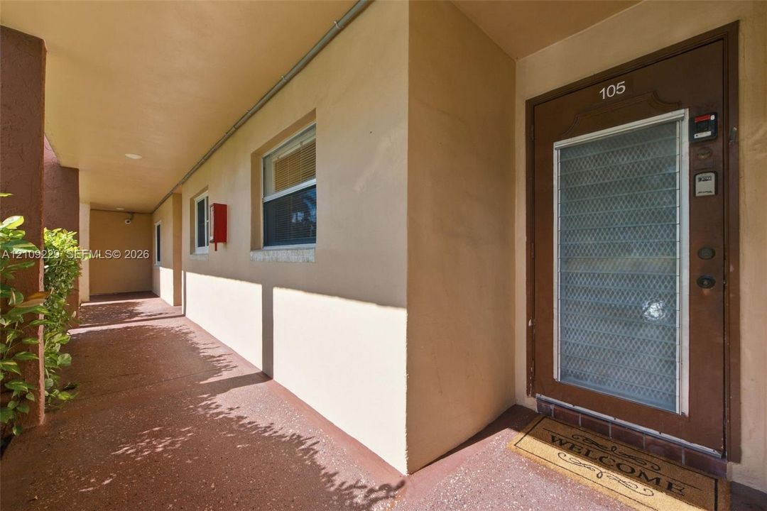 Covered exterior walkway with textured flooring, smooth stucco exterior, and a metal security door featuring an integrated glass panel