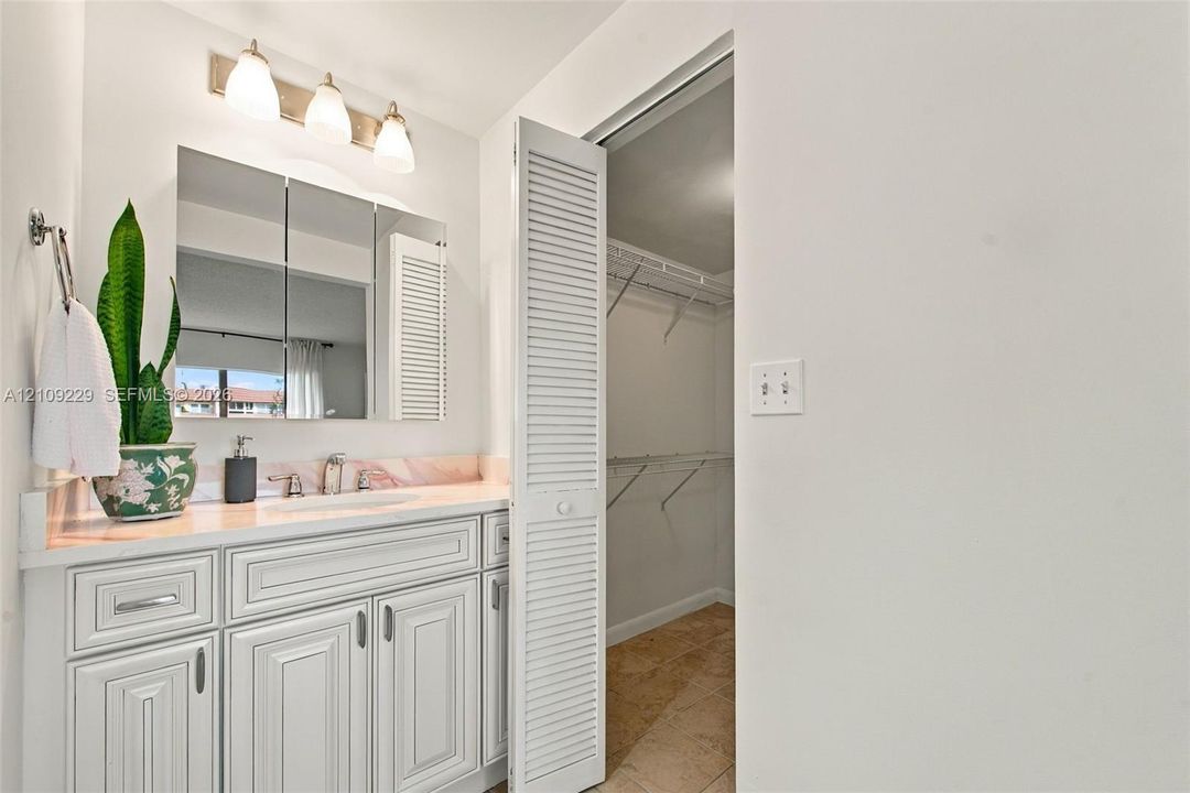 Vanity featuring white recessed-panel cabinetry, integrated stone-finish countertop with backsplash, three-light wall fixture, and tri-fold mirrored medicine cabinet, paired with a louvered-door walk-in closet
